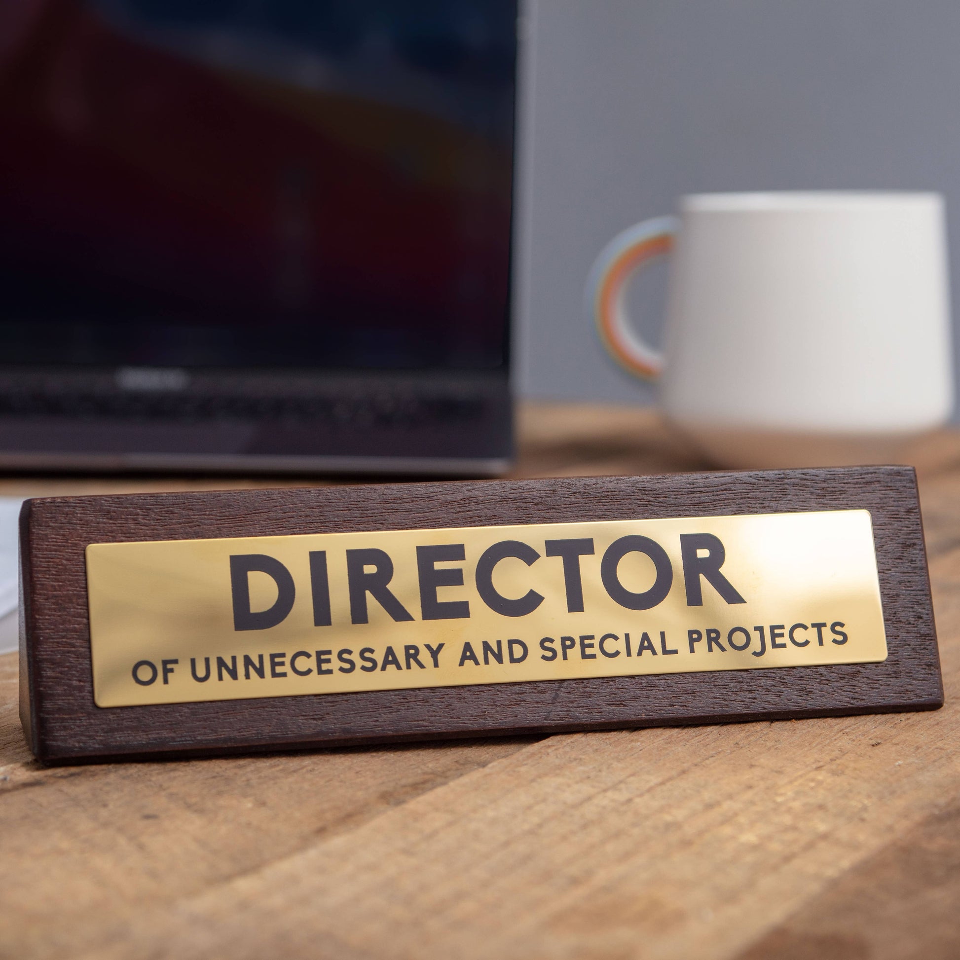 Wooden nameplate with 'DIRECTOR OF UNNECESSARY AND SPECIAL PROJECTS' on a desk with a laptop and mug in the background.