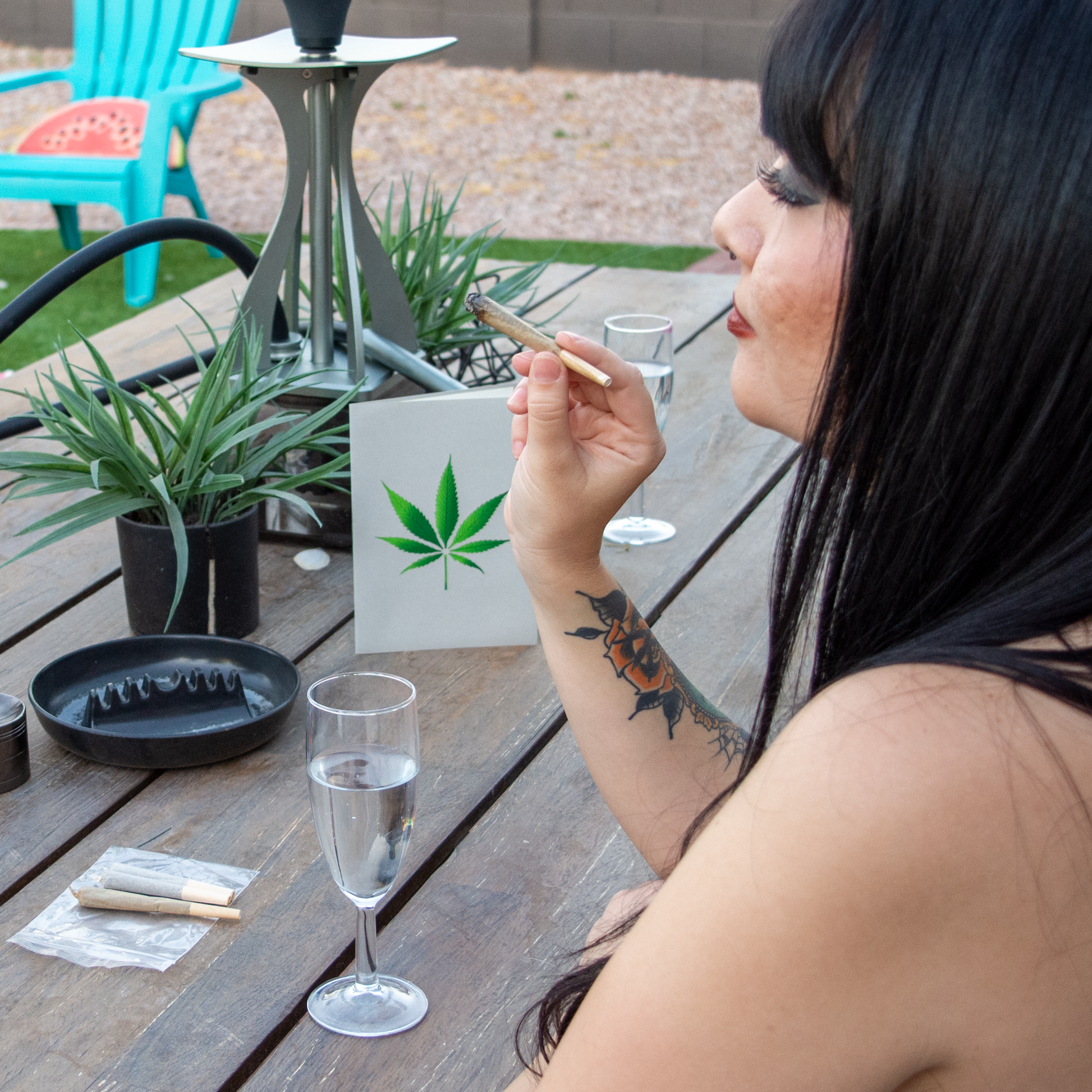 Woman smoking a cigarette outdoors with a cannabis leaf sign and drink on a table.