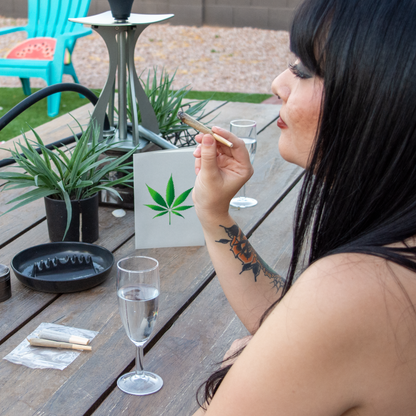 Woman smoking a cigarette outdoors with a cannabis leaf sign and drink on a table.