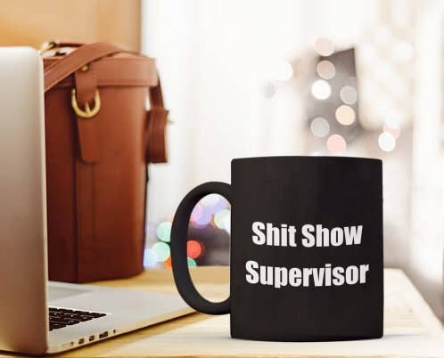 Black mug with 'Shit Show Supervisor' text on a desk with laptop and bag