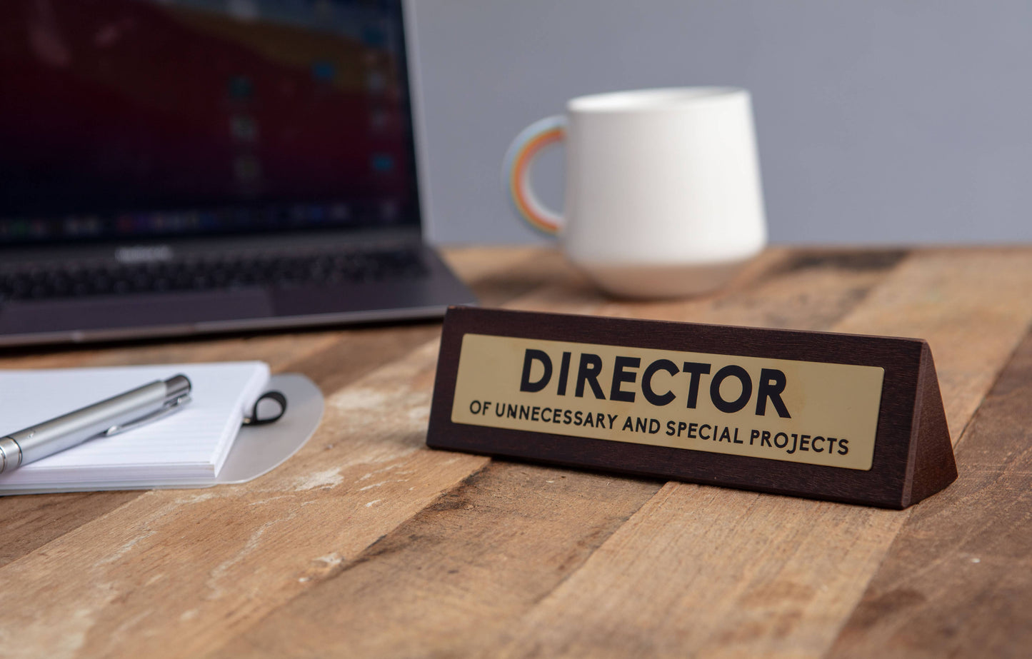 Desk with a nameplate labeled 'Director of Unnecessary and Special Projects' next to a laptop and mug.