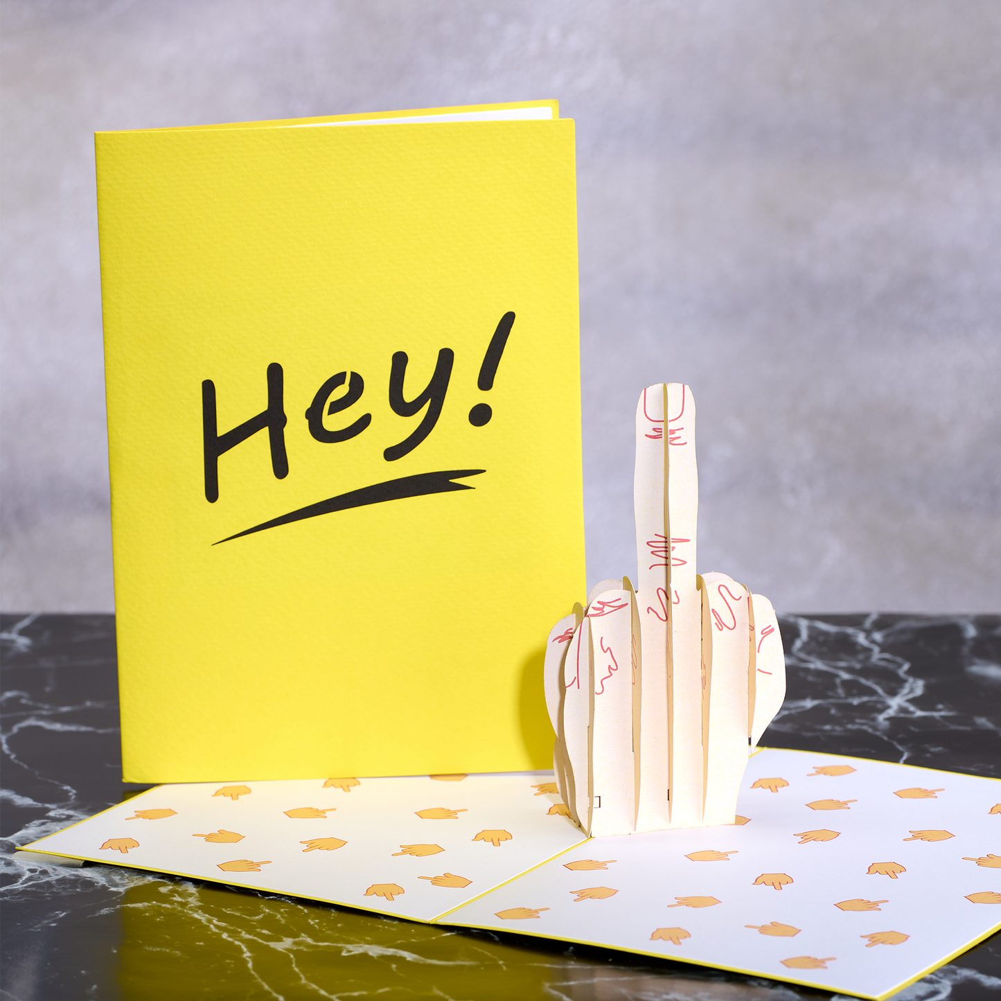 Yellow card with 'Hey!' text and a 3D hand gesture card on a gray surface.